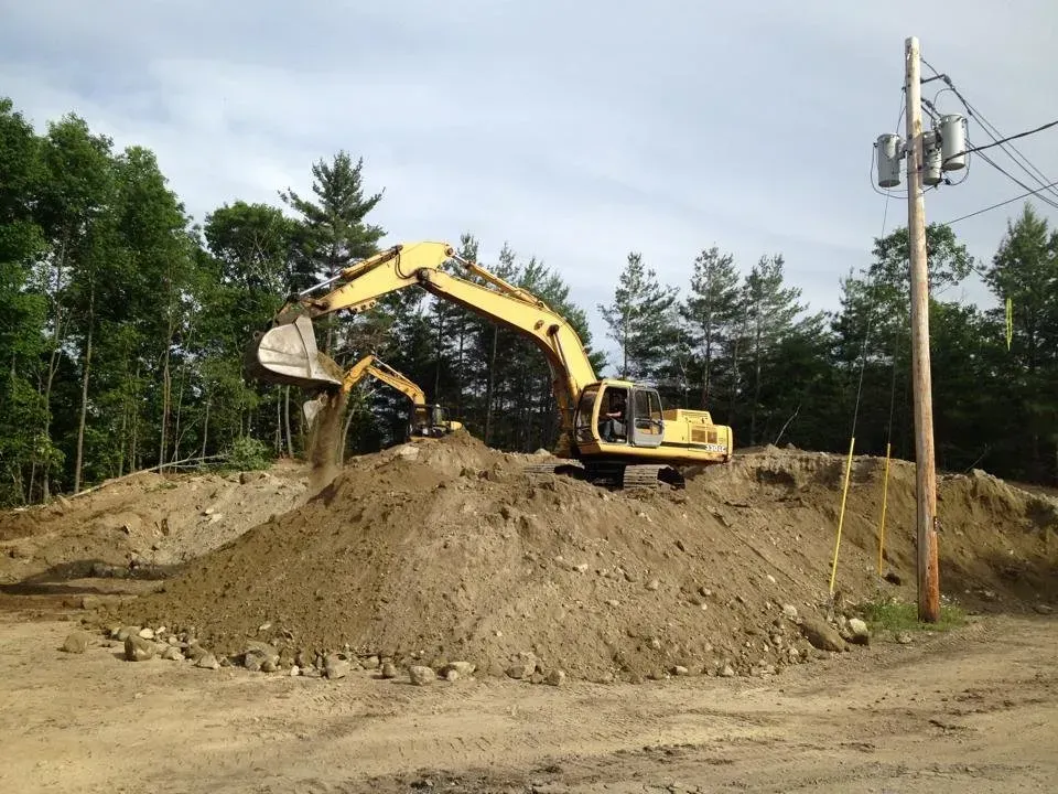 A yellow excavator works on a large dirt pile in a wooded area near a utility pole.