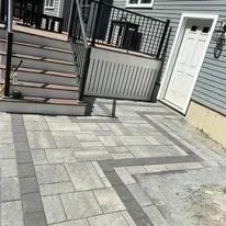 A gray, multi-toned stone patio with a dark border leads to a set of stairs and a white door on a gray-sided house.