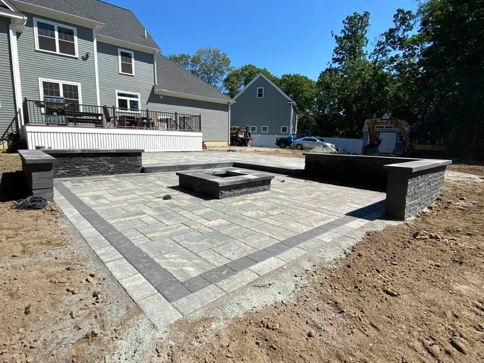 A newly installed stone patio with a central square fire pit and low retaining walls, situated behind a grey house.
