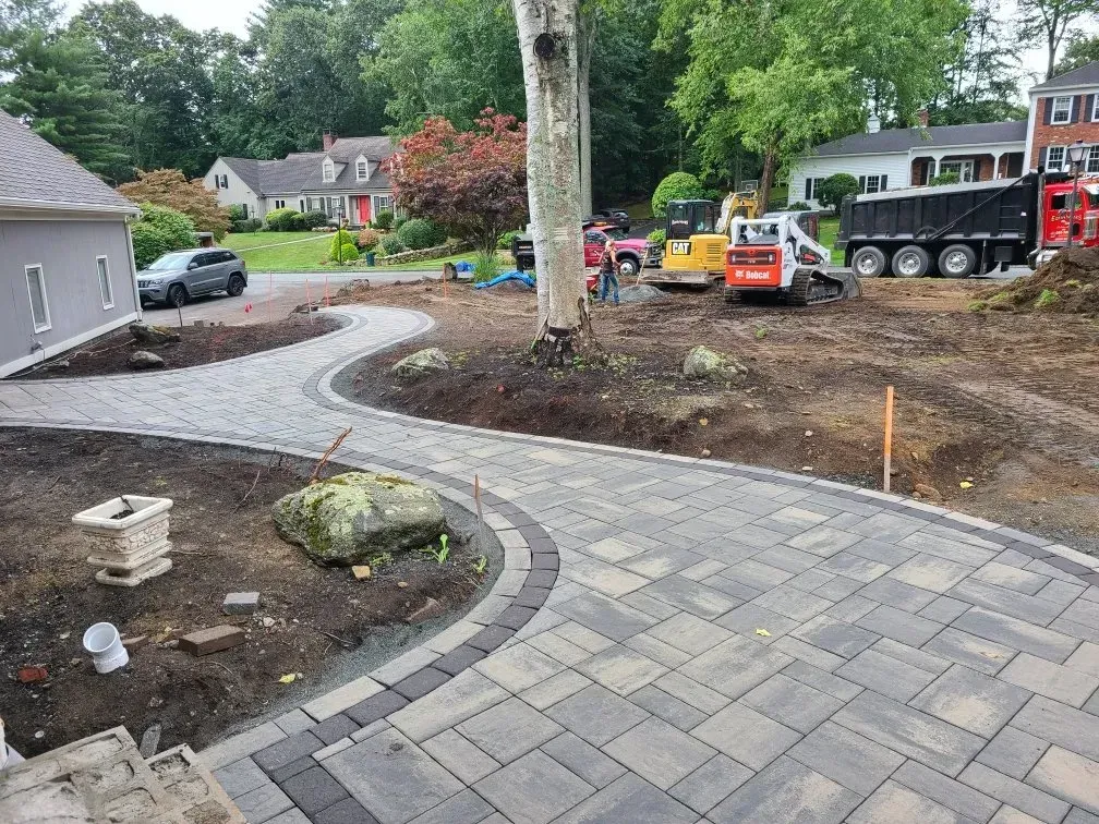 A newly installed gray paver walkway leads toward a house under construction with landscaping equipment in the background.