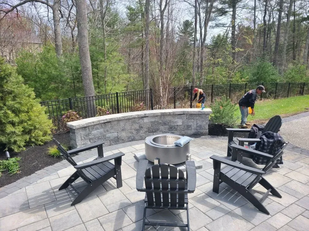A patio with a circular fire pit and three black chairs, with two people working in the background yard.