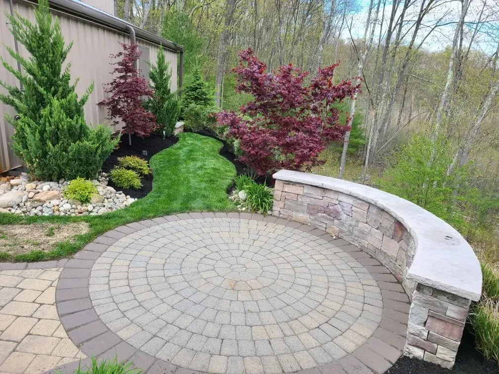 A circular stone patio features a curved stone seating wall, surrounded by landscaping with green and purple-leafed trees.