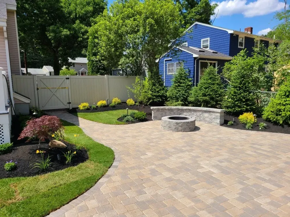 A paved patio with a circular stone fire pit and curved seating wall in a suburban backyard with a white fence.