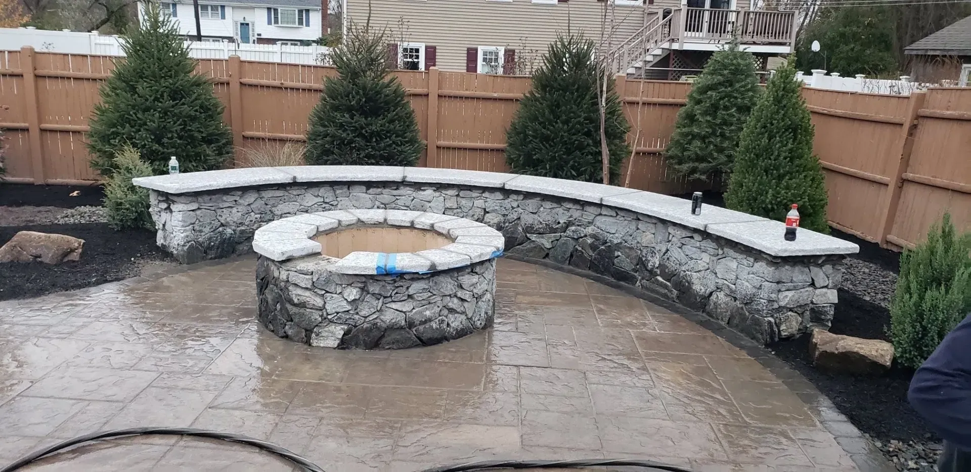 A stone fire pit surrounded by a curved stone seating wall on a paved patio in a fenced backyard with evergreen trees.