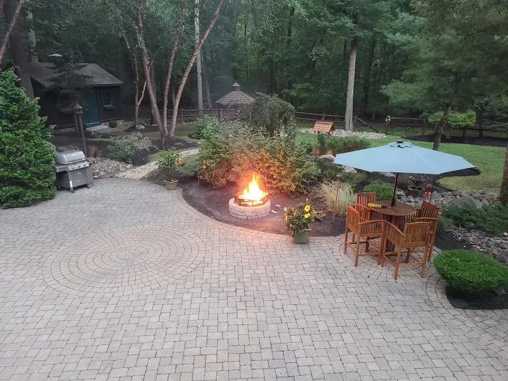 A stone patio with a lit fire pit and a table with an umbrella, set in a backyard surrounded by trees.