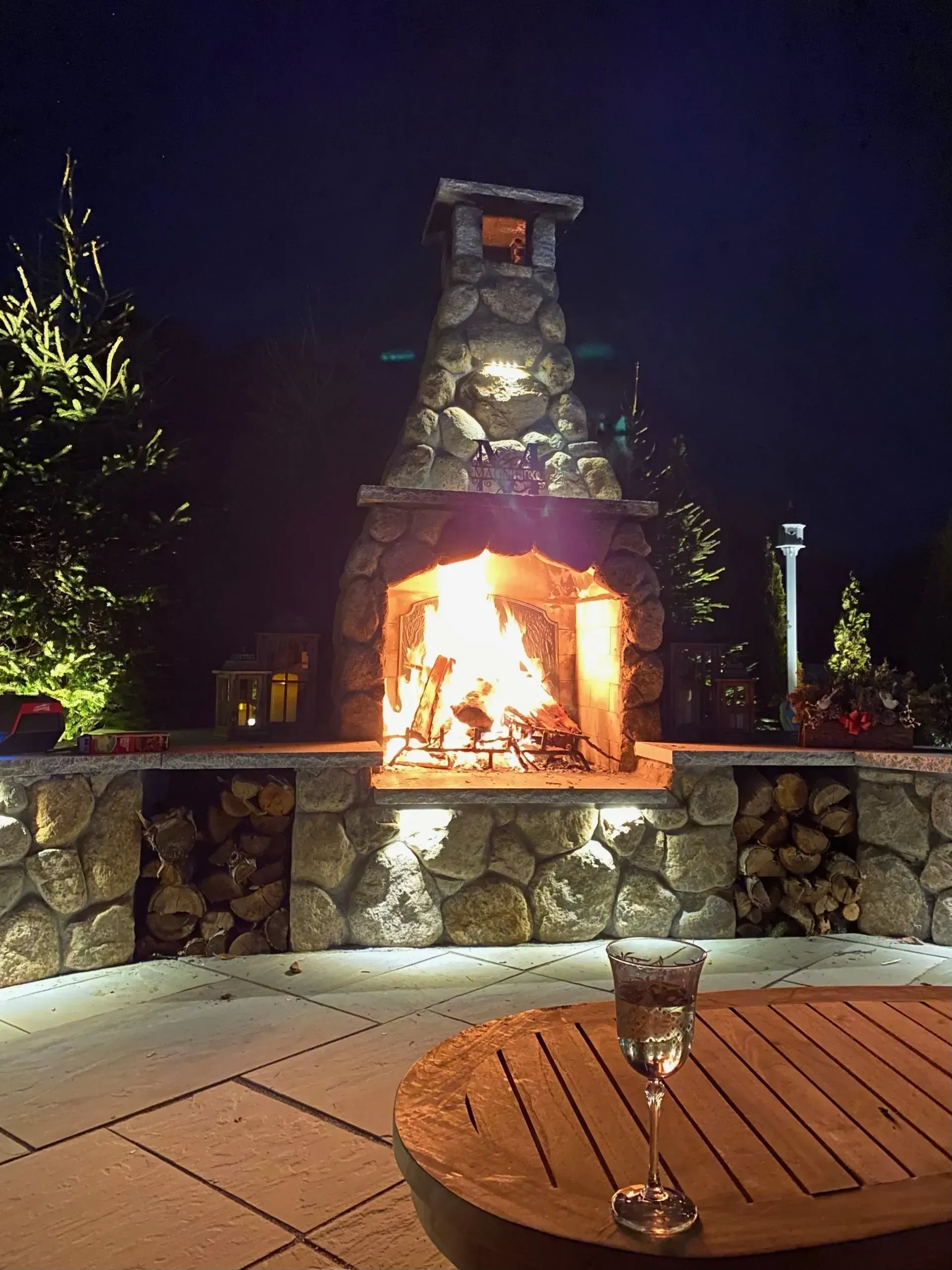 A cozy outdoor stone fireplace with a glowing fire, firewood storage, and a glass on a wooden table at night.