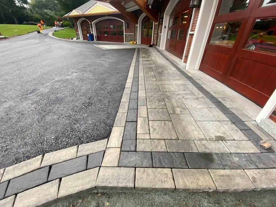 A paved walkway with a light stone center and dark stone border runs alongside the brown wooden doors of a building.