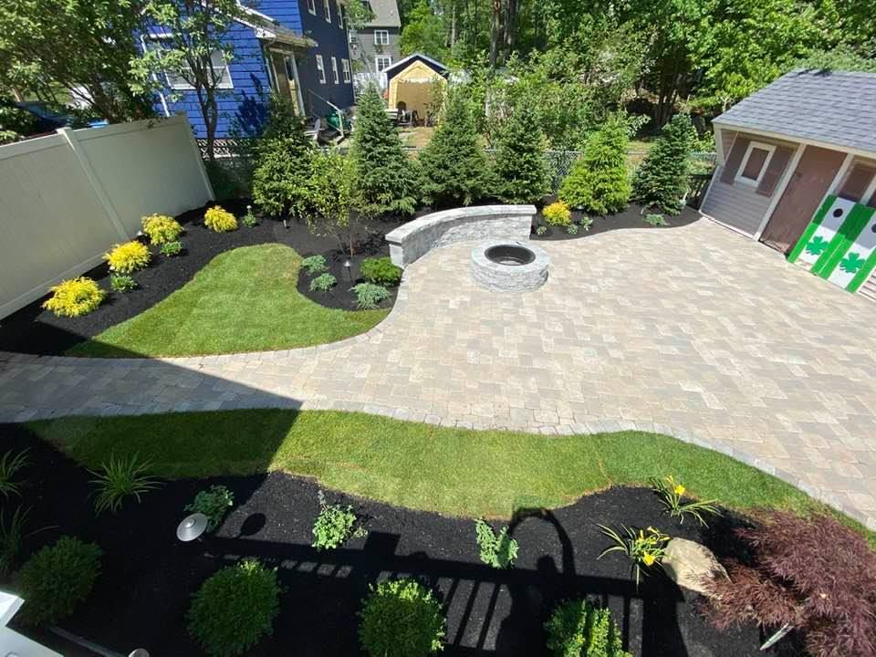 A landscaped backyard featuring a stone paver patio, a fire pit with a curved bench, lush green lawn, and garden beds.