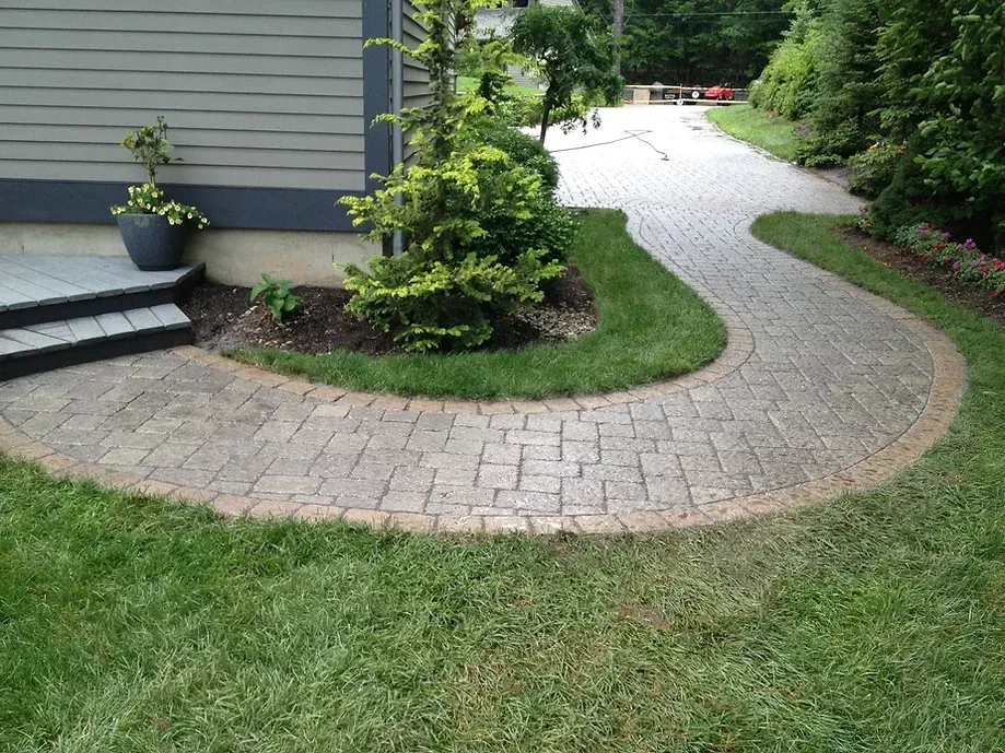 A curved stone paver walkway leads from concrete steps near a house wall across a green lawn toward a paved driveway.
