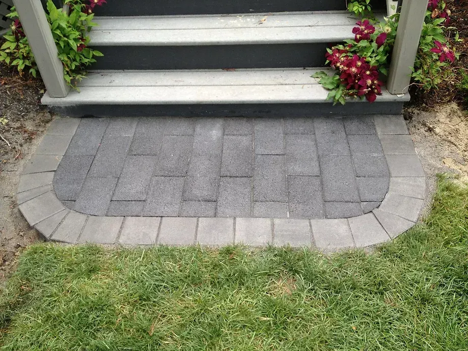 A small, semi-circular gray brick patio sits at the base of wooden stairs, flanked by greenery and red flowers.