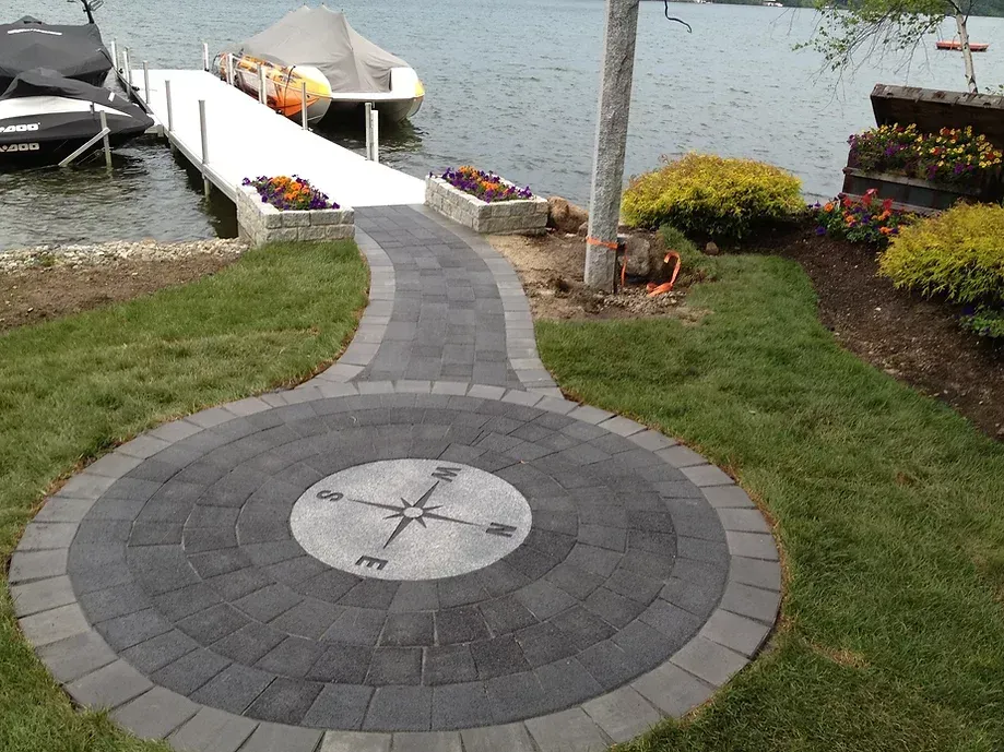 A stone patio with a compass rose inlay leads to a walkway ending at a lake dock with boats.