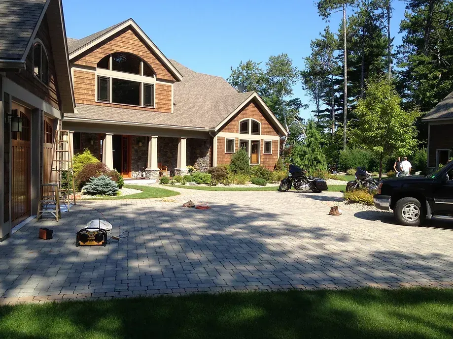 A large, multi-story brown shingle house with a stone-paved driveway, motorcycles, and a parked black truck.