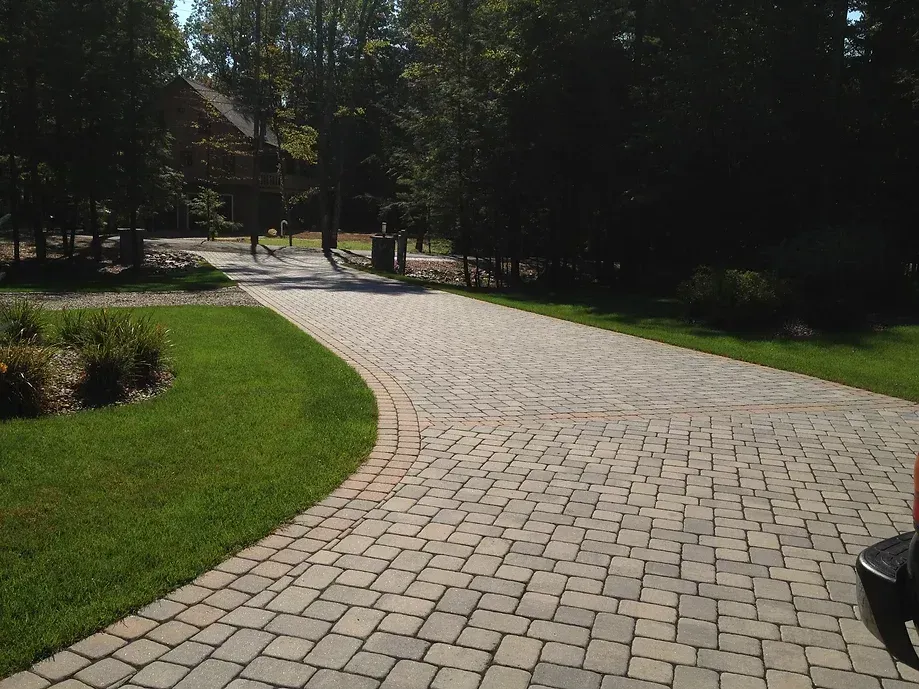 A curved driveway paved with light gray and tan stone blocks, bordered by green grass and surrounded by trees.