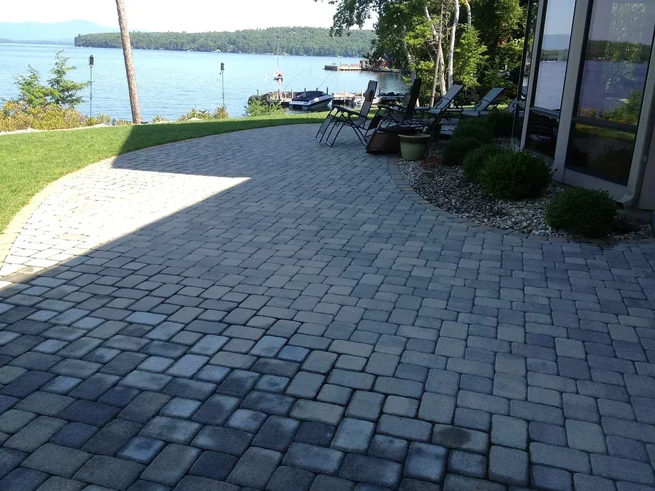 A stone patio overlooking a lake with lawn chairs, landscaping, and a view of trees and distant hills on a sunny day.
