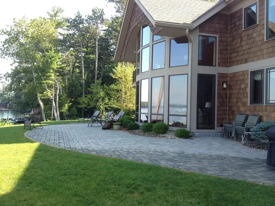 A modern house with large windows and shingled siding overlooks a stone patio with outdoor seating near a wooded lake.