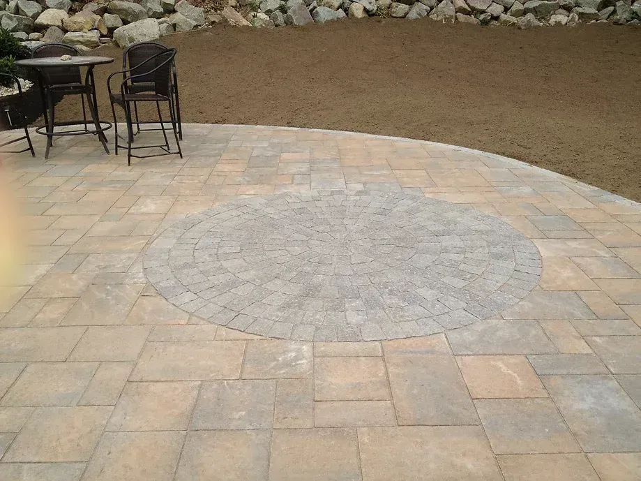 A patio made of tan and gray pavers with a circular accent pattern, featuring a small bistro table and chairs.