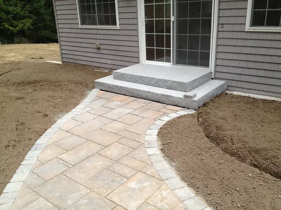 A stone walkway with a decorative border leads to gray concrete steps in front of a house with light gray siding.