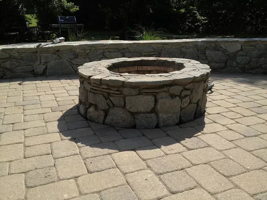 A circular stone fire pit sits on a light-colored paver patio in front of a low stone wall.