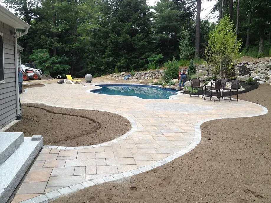 A newly installed backyard patio with light-colored pavers surrounding a curved in-ground pool near a house and woods.