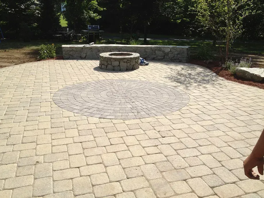 A stone patio featuring a circular paved inlay, a stone fire pit, and a curved stone seating wall in a backyard.