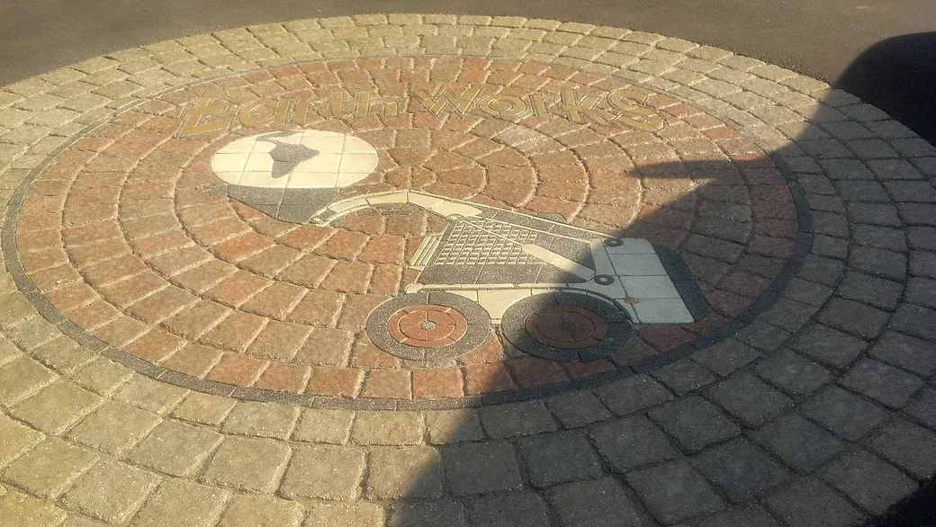 A circular stone patio features a mosaic of a map and a gear-like mechanism in grey and reddish-brown pavers.