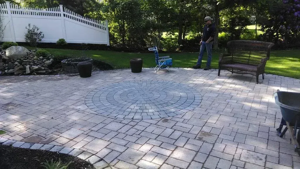 A stone patio featuring a circular pattern in the center, a person standing near a bench, and a white fence in the back.