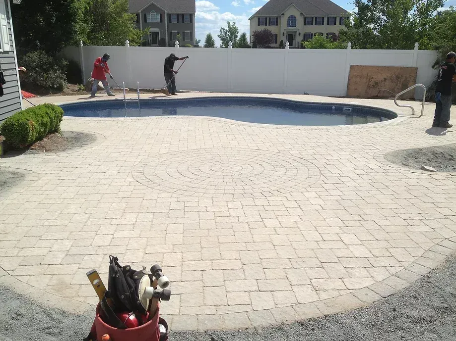 Three workers install light-colored pavers around a backyard swimming pool in front of a white privacy fence.