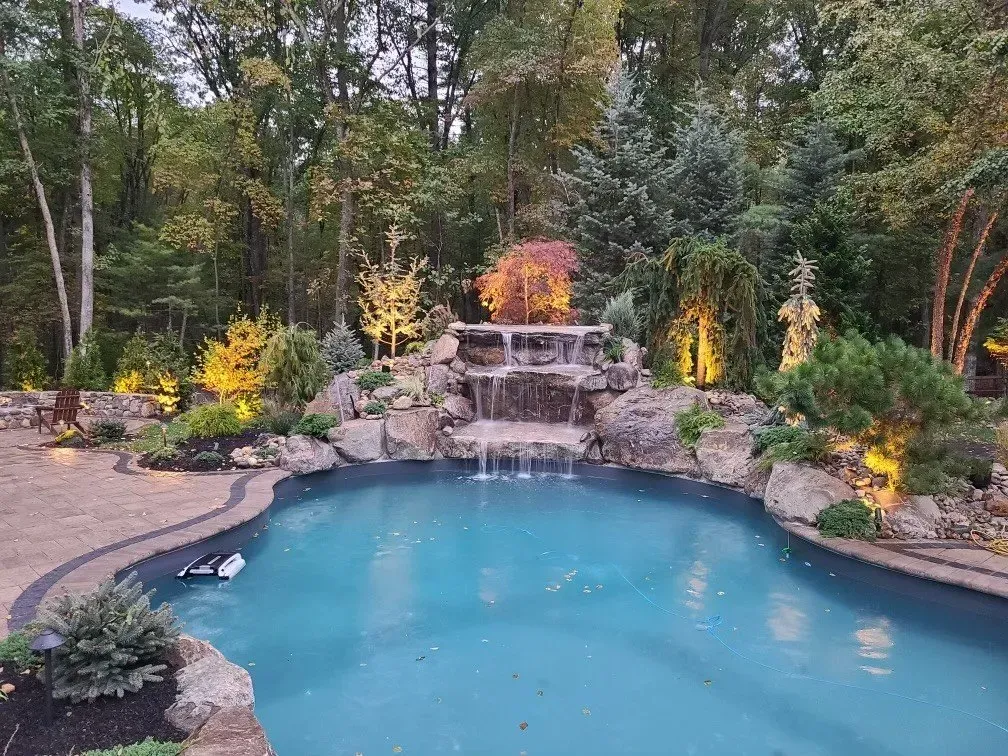 An illuminated backyard swimming pool with a rock waterfall feature surrounded by lush trees at dusk.