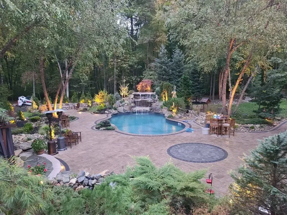 A backyard oasis with a blue swimming pool, stone patio, waterfall feature, and lush trees illuminated by landscape lights.