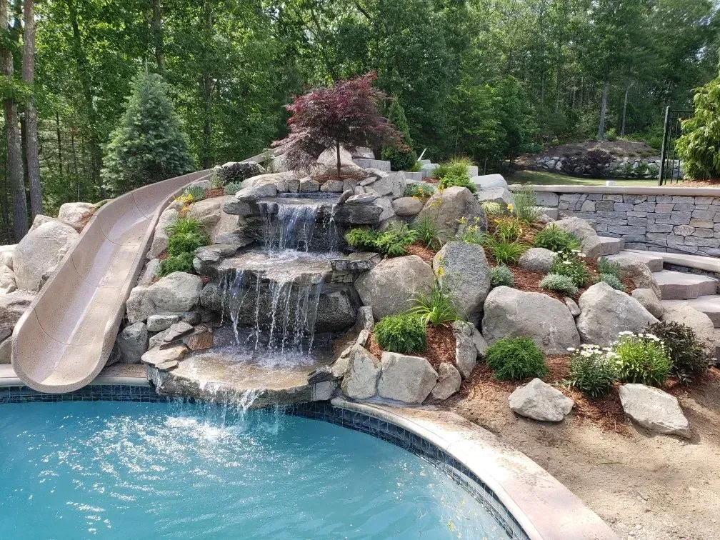 A backyard swimming pool with a multi-tiered rock waterfall and an integrated slide on the left.
