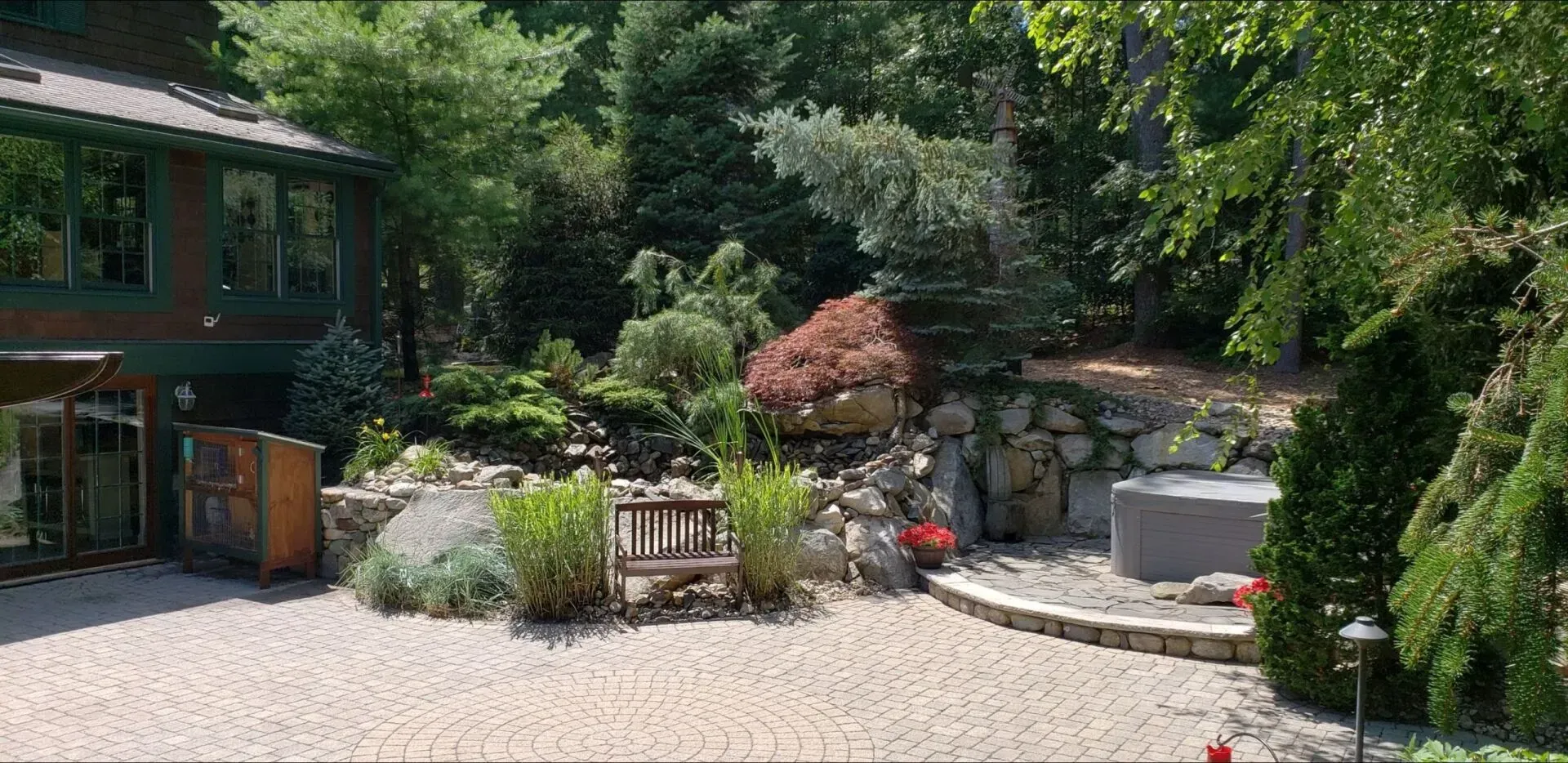 A stone-paved patio featuring a wooden bench surrounded by rock landscaping, lush greenery, and a house exterior.