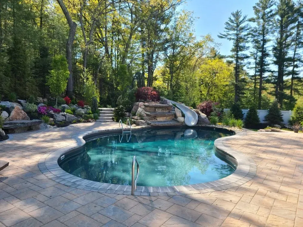 A kidney-shaped swimming pool with a stone slide and patio, surrounded by lush trees and a landscaped garden.