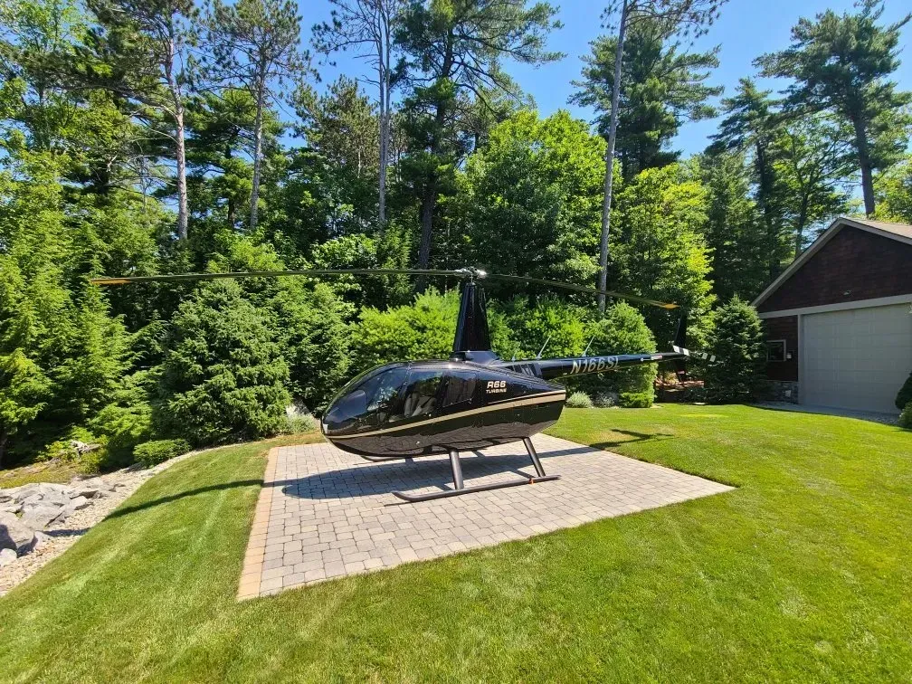 A sleek black helicopter parked on a stone pad in a grassy yard, surrounded by lush green trees and a building.