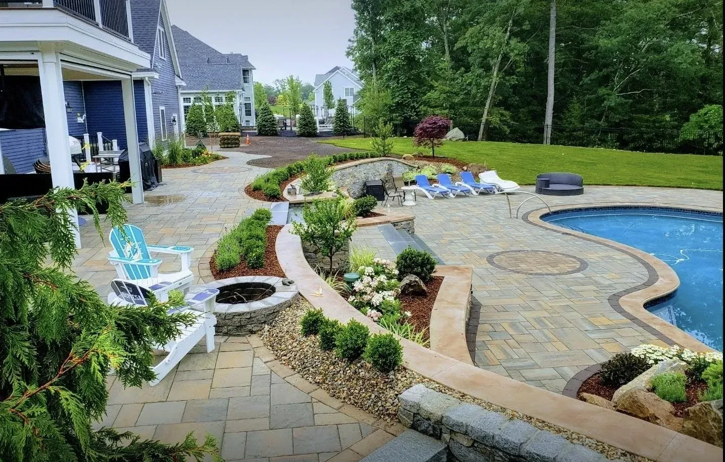 A backyard patio with stone pavers, a swimming pool, a fire pit area, and landscaped garden beds near a house.