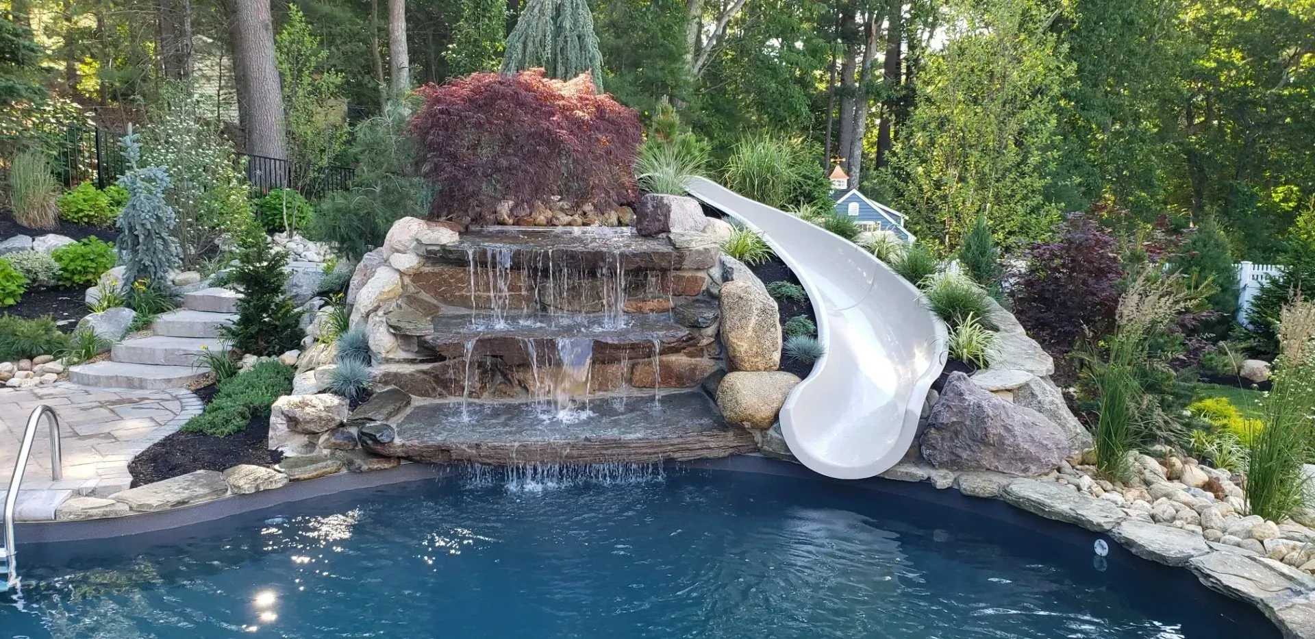 A multi-tiered stone waterfall feature emptying into a swimming pool with a curved white water slide alongside it.