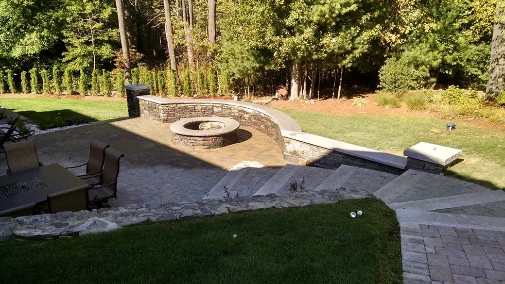 A backyard patio with a stone fire pit, seating area, and wide steps leading to a lawn with trees in the background.