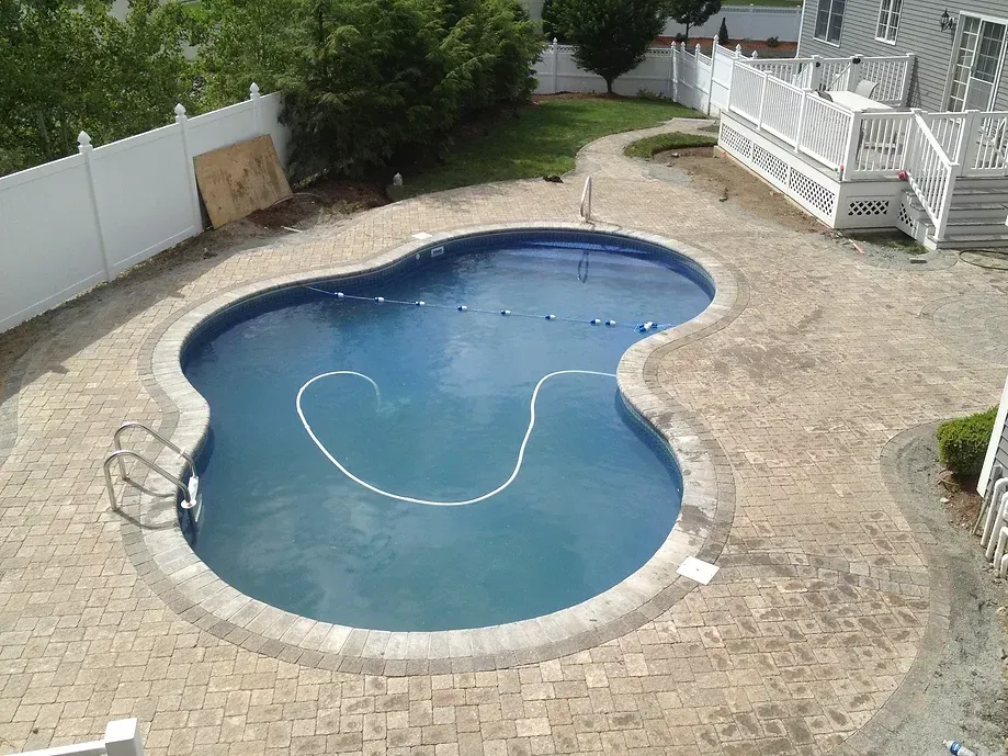 An elevated view of a kidney-shaped backyard swimming pool surrounded by tan stone pavers, a white fence, and a deck.
