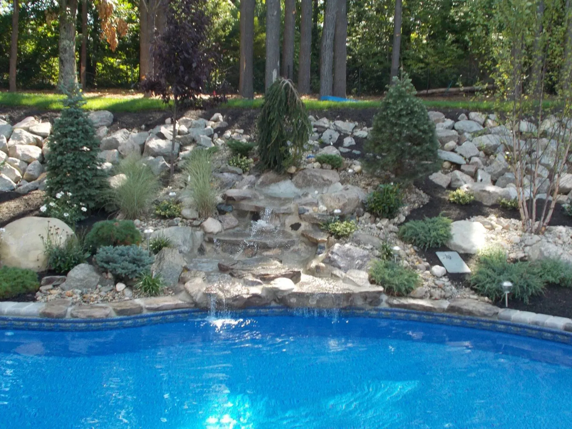 A blue swimming pool features a stone waterfall cascade set into a landscaped hillside with boulders and evergreen trees.