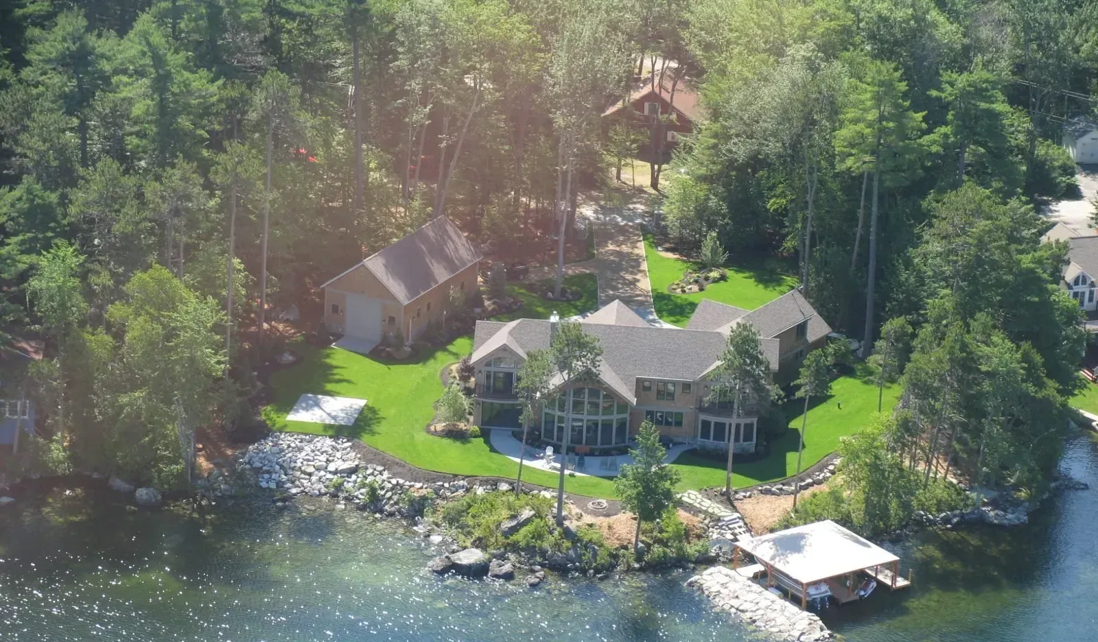 Aerial view of a large waterfront home with a grassy lawn, detached garage, and private dock surrounded by trees.