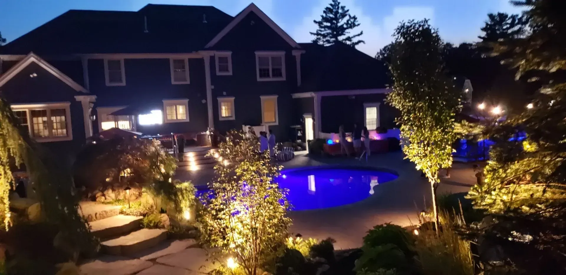 A luxurious house with a glowing blue swimming pool at night, surrounded by illuminated trees and stone steps.