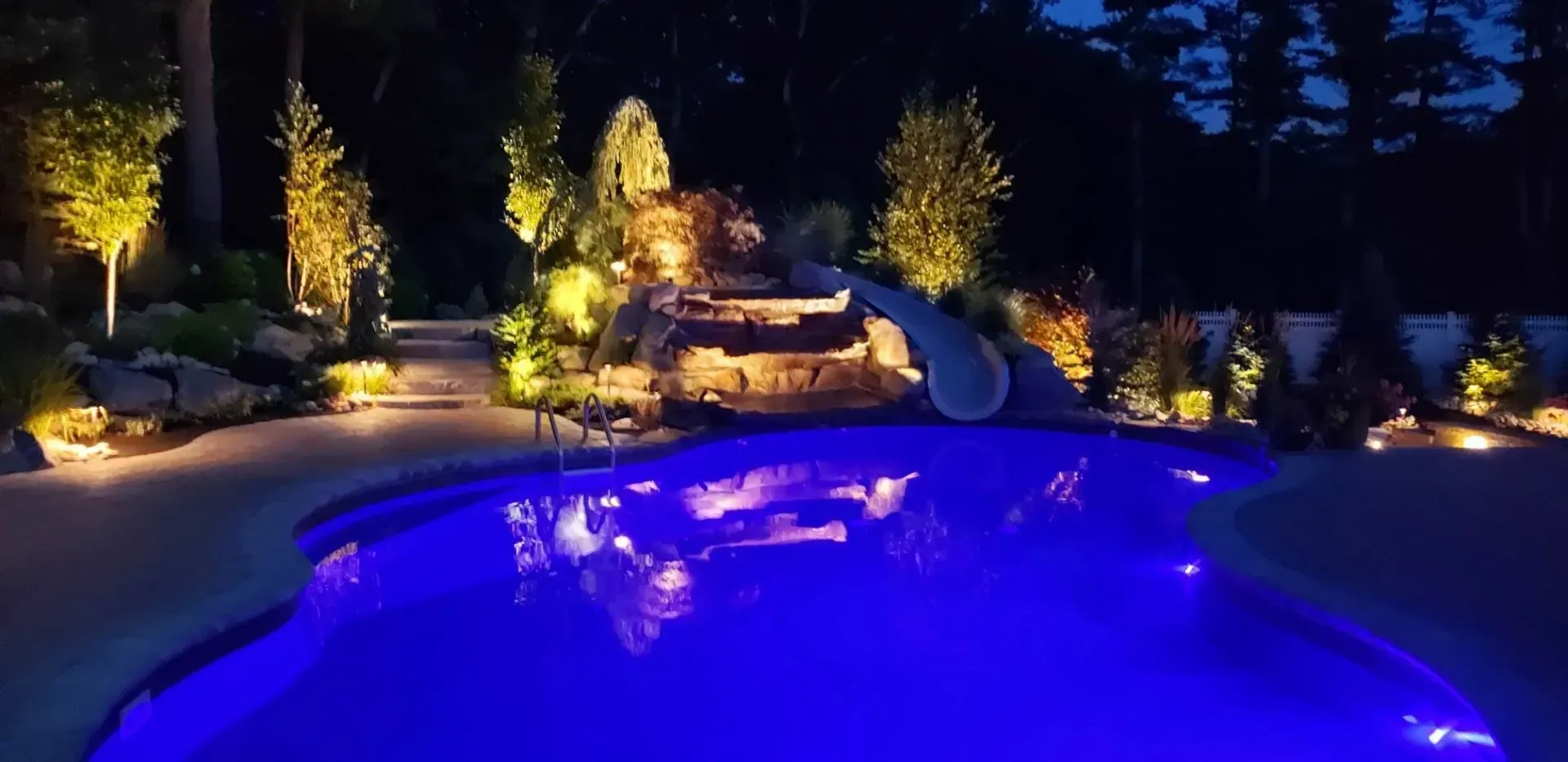 A glowing blue swimming pool at night with a rock waterfall, slide, and illuminated landscaping in the background.