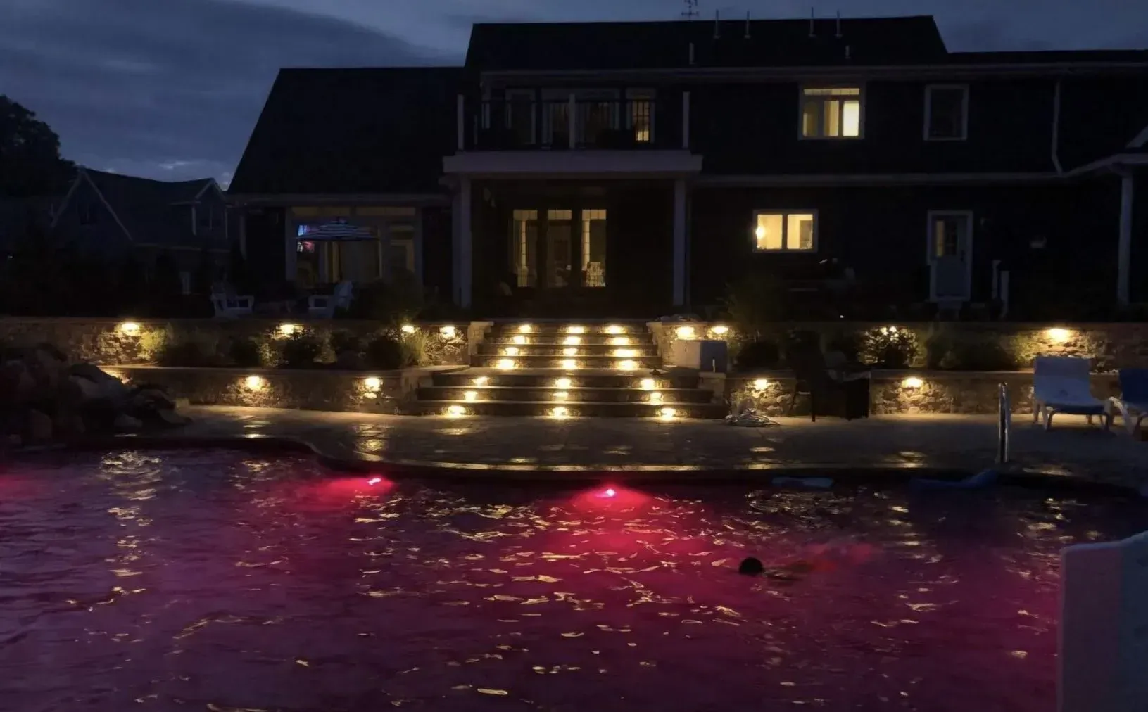 A backyard pool lit with pink underwater lights at night, featuring a stone staircase and patio illuminated by soft lamps.