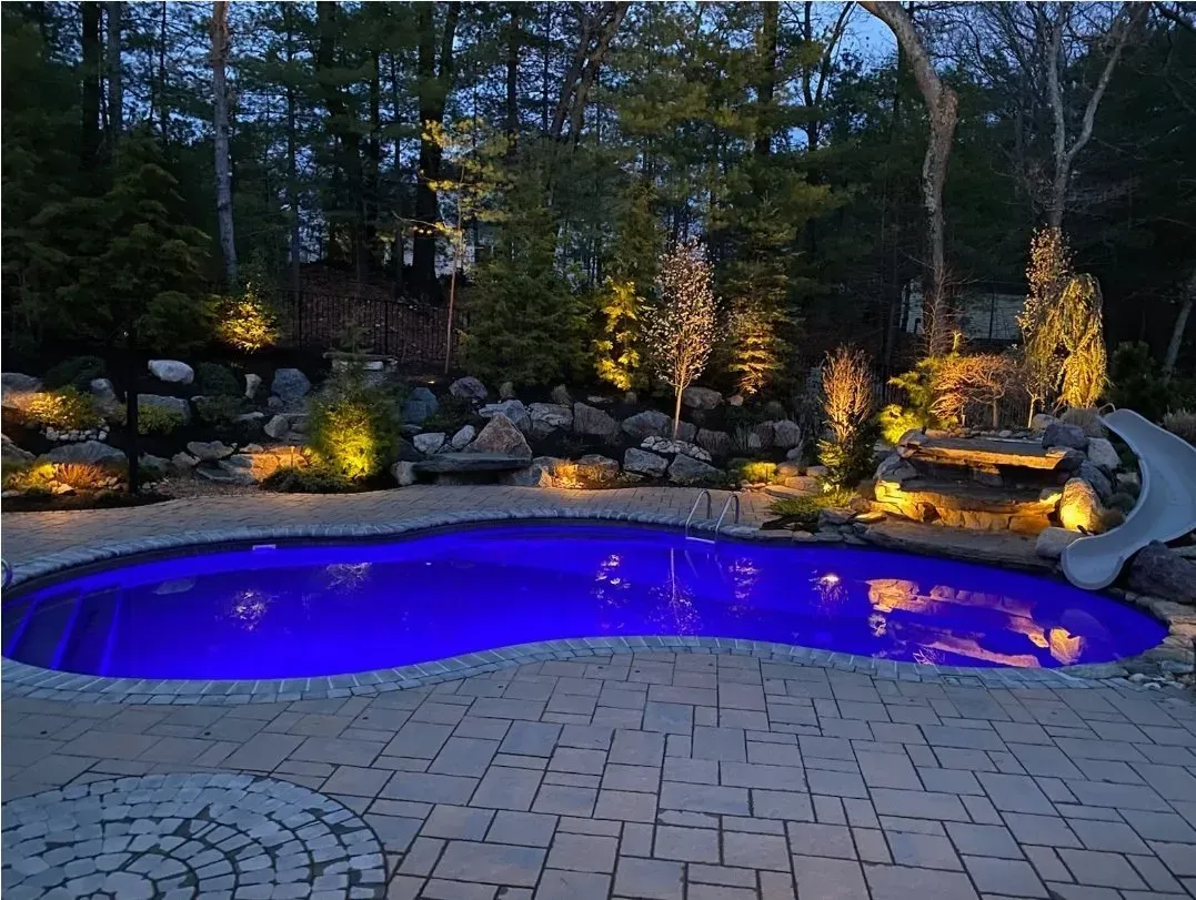 A blue-lit swimming pool surrounded by a stone patio and landscaped garden with outdoor lighting at dusk.