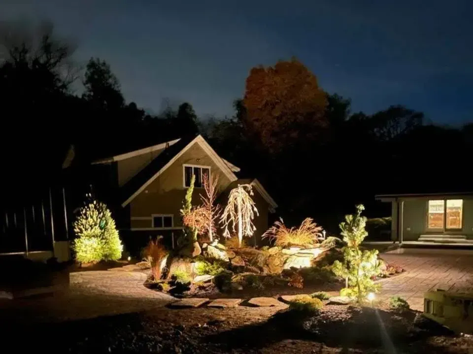 A home exterior at night featuring illuminated landscaping, trees, and stone walkways with warm accent lighting.