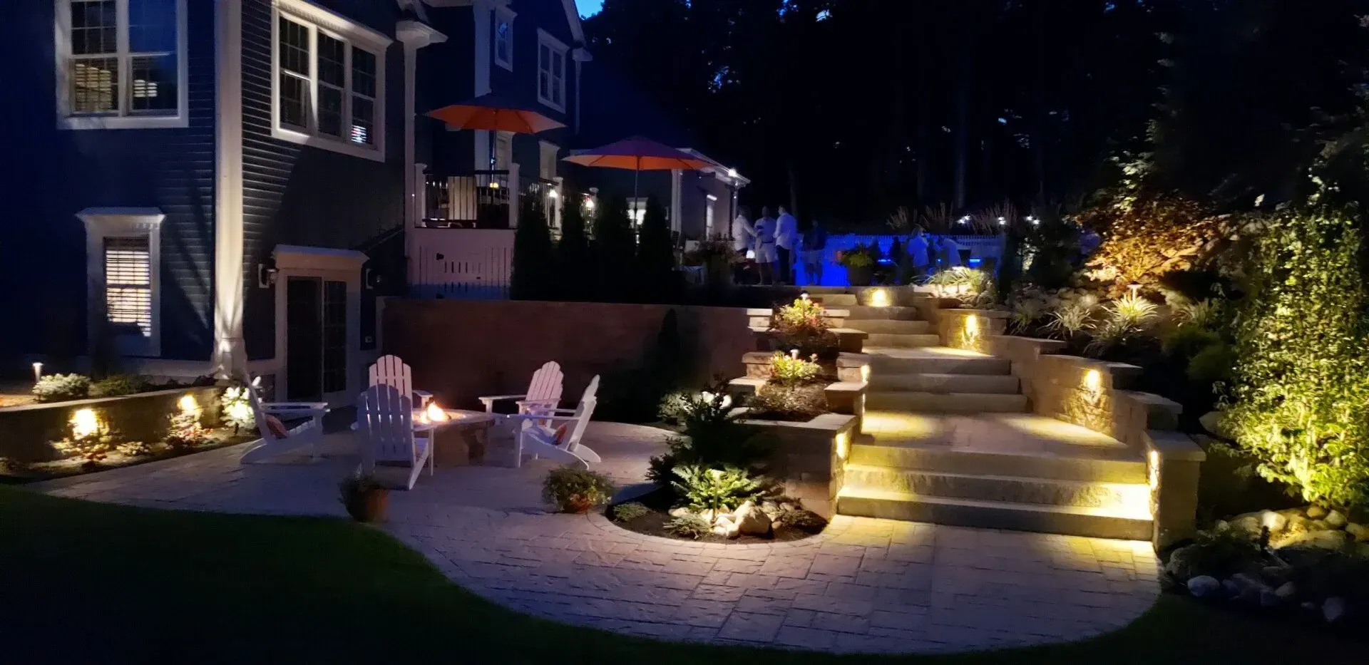 An illuminated stone patio with a fire pit, Adirondack chairs, and lit steps leading to a backyard garden at night.
