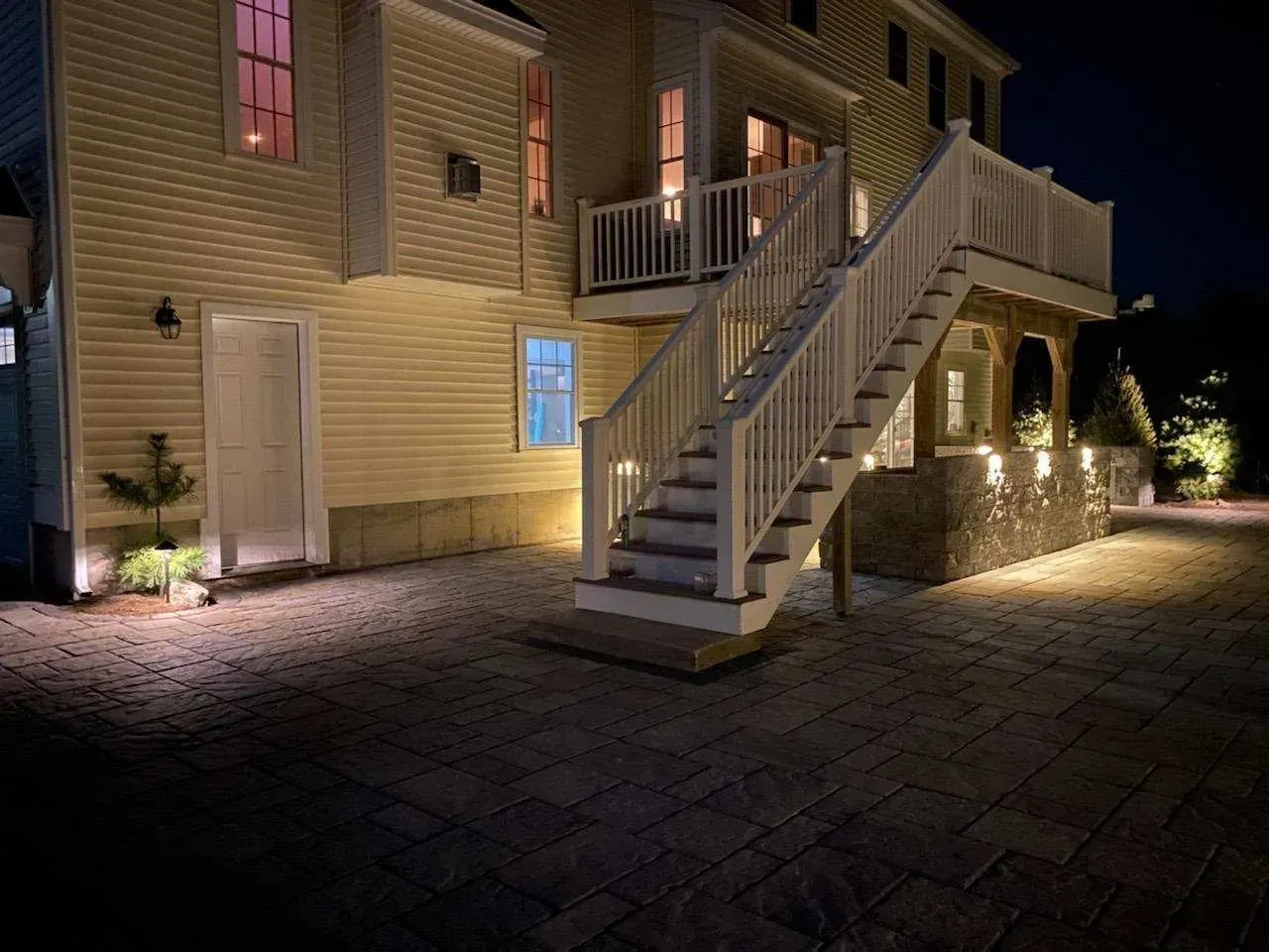 A yellow house at night with a stone patio, outdoor staircase, and warm accent lighting on the walls and ground.