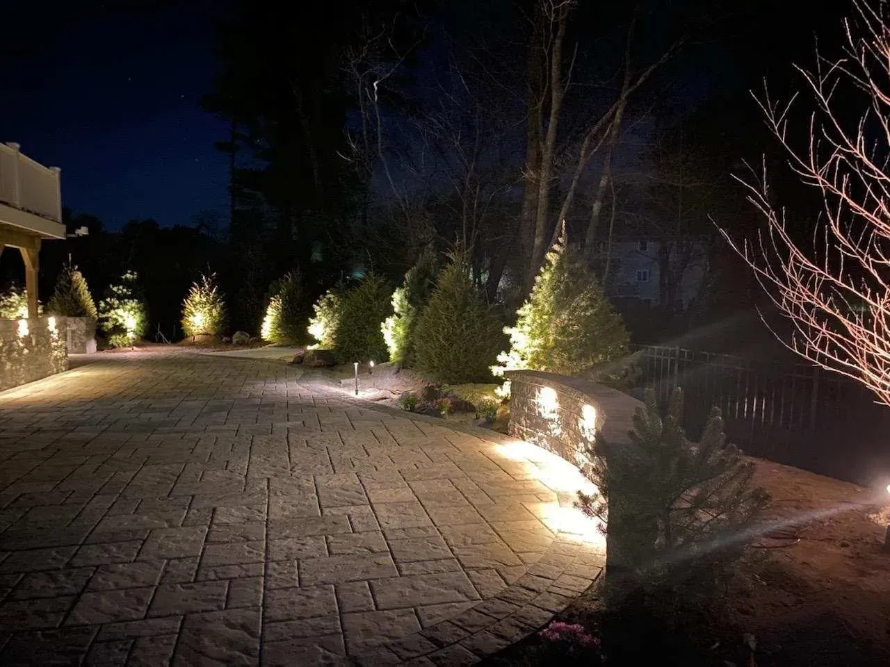 A stone patio at night illuminated by warm landscape lighting, highlighting a row of evergreen trees and a retaining wall.