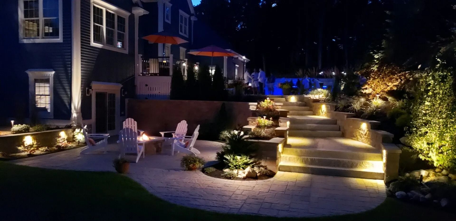 Night view of a backyard patio with a stone fire pit, Adirondack chairs, and stone steps illuminated by landscape lights.