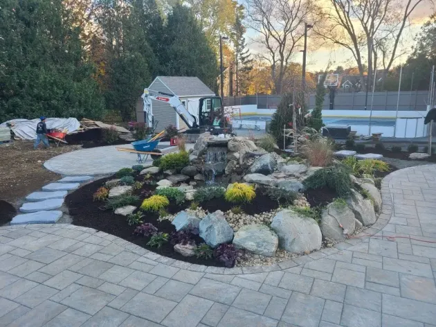A landscaped backyard with stone pavers, a central rock waterfall, garden beds, and an in-ground pool in the distance.