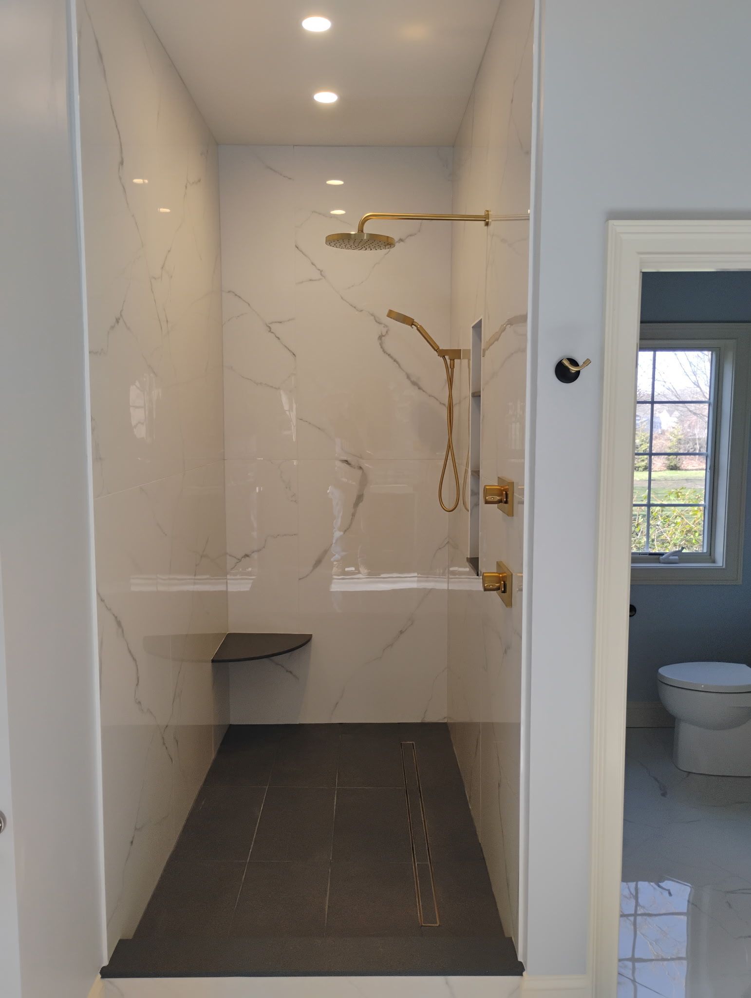Narrow tiled shower with built-in bench, marble-look walls, and a partially visible bathroom doorway.